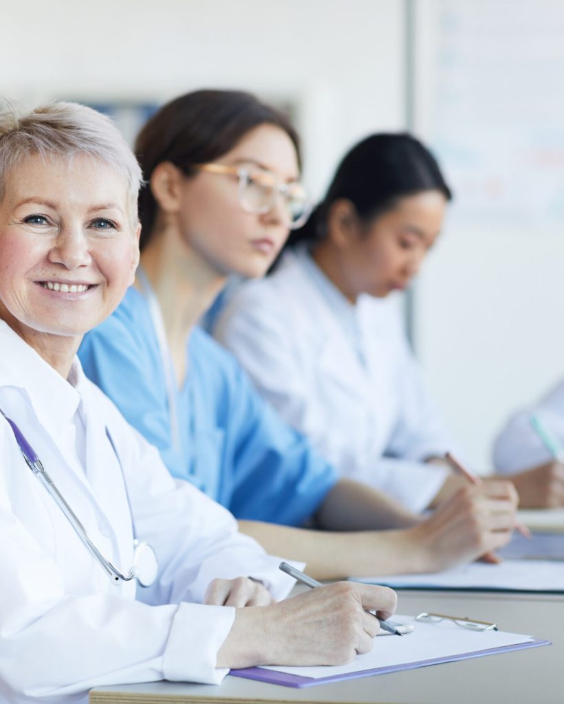 Portrait of mature female doctor smiling at camera while sitting at table during medical council or conference in clinic, copy space